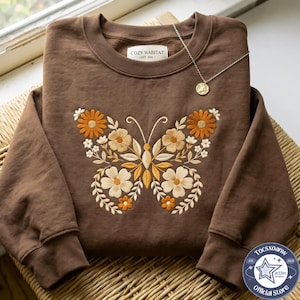May include: A brown crewneck sweatshirt featuring an embroidered butterfly design composed of various orange, cream, and tan flowers and leaves. The inner neck label reads COZY HABITAT EST. 2021. A thin gold chain necklace rests on the fabric.