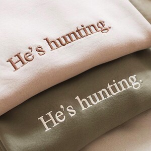 May include: Two folded sweatshirts, one light pink and one olive green, are stacked. The phrase "He's hunting." is embroidered on each in contrasting colors. The light pink sweatshirt has brown embroidery, while the olive green one has white.