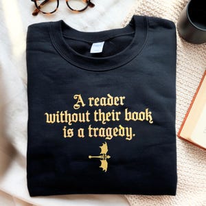 May include: Black sweatshirt with gold text reading "A reader without their book is a tragedy." A gold dragon and sword illustration is below the text. The folded sweatshirt is on a cream-coloured surface.