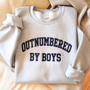 May include: A light gray sweatshirt with the text "OUTNUMBERED BY BOYS" in navy blue lettering. The sweatshirt has a crew neck and long sleeves, with the sleeves folded up. The garment is made of a soft material.
