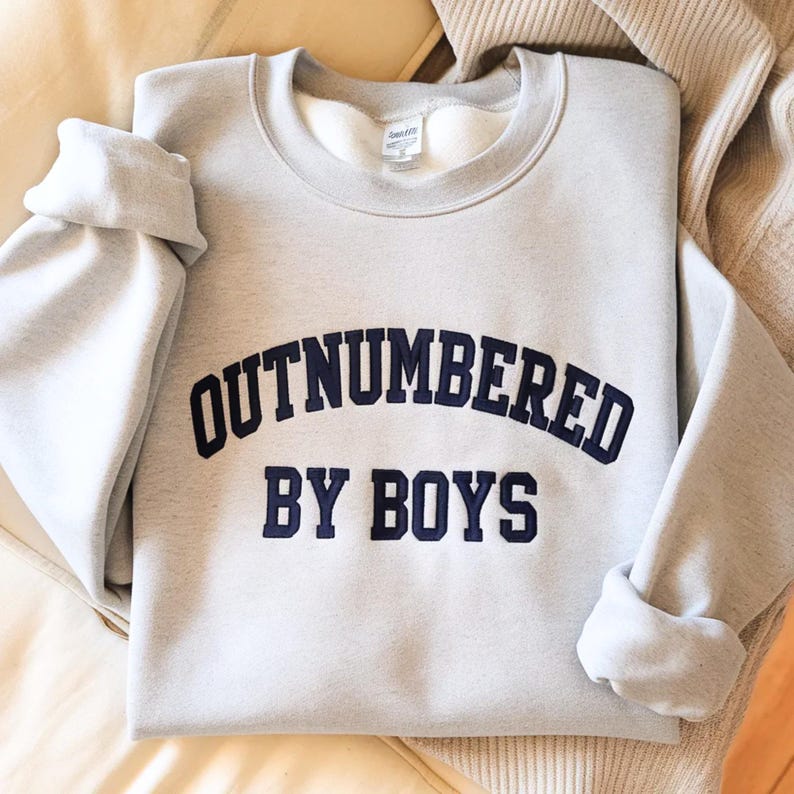 May include: A light gray sweatshirt with the text "OUTNUMBERED BY BOYS" in navy blue lettering. The sweatshirt has a crew neck and long sleeves, with the sleeves folded up. The garment is made of a soft material.