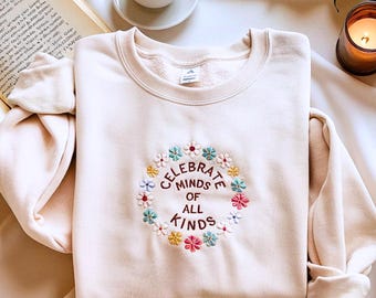 May include: A cream-colored sweatshirt with a crew neck and long sleeves. The front features an embroidered floral wreath with the words "CELEBRATE MINDS OF ALL KINDS" in a circular design. The sweatshirt is folded on a white surface.