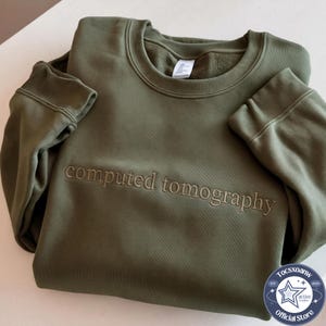 May include: An olive green crewneck sweatshirt featuring the text computed tomography embroidered in a matching thread color across the chest. The garment has long sleeves with ribbed cuffs and a ribbed neckline, with a white interior label.