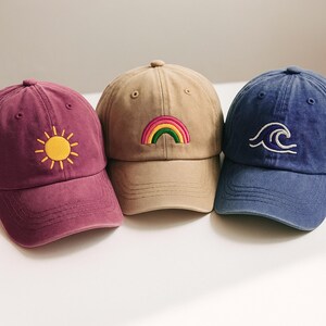 May include: Three baseball caps in burgundy, beige, and blue. The burgundy cap has a yellow sun embroidered on the front. The beige cap features a rainbow, and the blue cap has a white wave design. All caps have a curved peak.