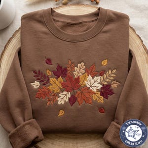 May include: A brown crewneck sweatshirt featuring an embroidered design of various autumn leaves in shades of red, orange, yellow, and cream. The leaves are arranged in a central cluster on the chest, with some individual leaves scattered.