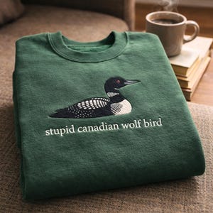 May include: A dark green sweatshirt featuring an embroidered image of a black and white bird with the text "stupid canadian wolf bird" in white. The sweatshirt is folded and resting on a brown surface.