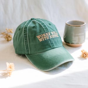 May include: A green baseball cap with the text "WORKS WELL WITH COFFEE" in a light brown colour. The cap is made of a soft material and has a curved peak. A ceramic mug is in the background.