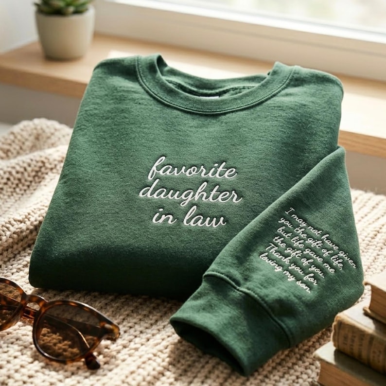 May include: A forest green crewneck sweatshirt featuring white embroidered text on the chest that reads favorite daughter in law. The sleeve includes additional white embroidered text that reads I may not have given you the gift of life, but life gave me the gift of you. Thank you for loving my son.