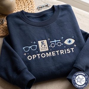 May include: A navy blue crewneck sweatshirt featuring embroidered designs of light blue glasses, an eye chart, a phoropter, and an eye. The word OPTOMETRIST is embroidered in cream-colored lettering below the icons, accented by small stars.