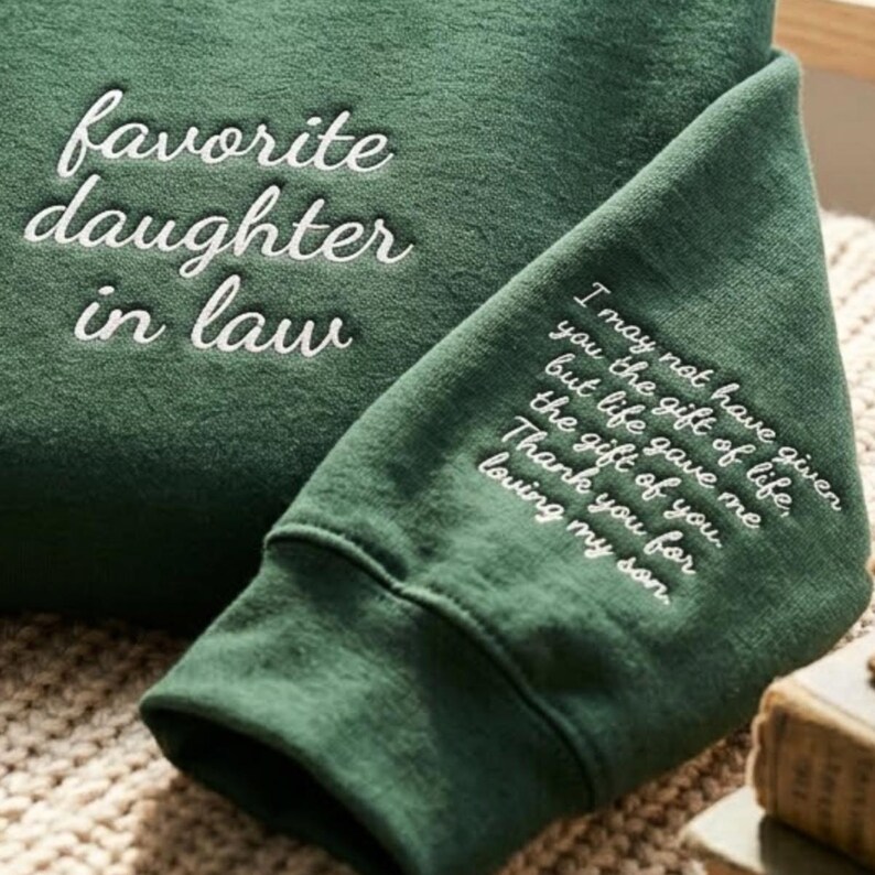 May include: A dark green sweatshirt featuring white embroidered text on the chest that reads favorite daughter in law. The sleeve includes additional white embroidered text that reads I may not have given you the gift of life, but life gave me the gift of you. Thank you for loving my son.