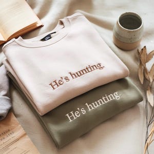May include: Two sweatshirts, one beige and one olive green, are stacked. The beige sweatshirt has the embroidered phrase "He's hunting" in a brown thread. The olive green sweatshirt has the same phrase embroidered in white.