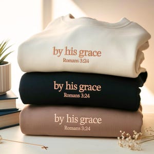 May include: A stack of three sweatshirts in cream, black, and taupe. Each sweatshirt has the embroidered text "by his grace" and "Romans 3:24" in a matching rose-gold color. The sweatshirts are neatly folded and stacked on top of each other.