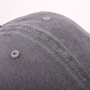 May include: Close-up of a gray baseball cap. The cap is made of a textured fabric and features a stitched seam and a small, embroidered ventilation hole. The cap's design is simple and classic.