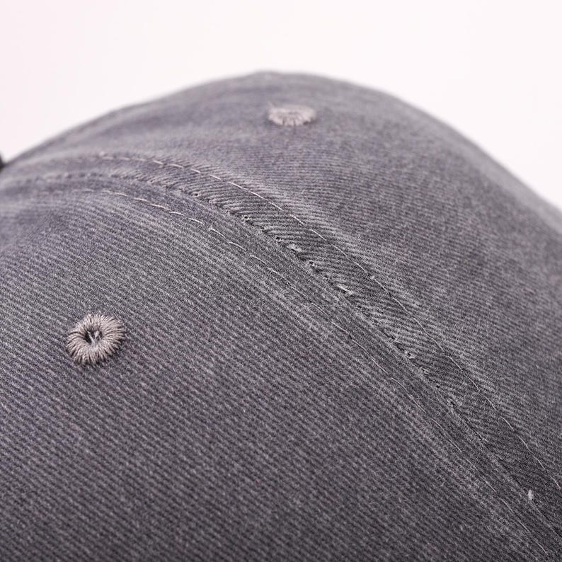 May include: Close-up of a gray baseball cap. The cap is made of a textured fabric and features a stitched seam and a small, embroidered ventilation hole. The cap's design is simple and classic.