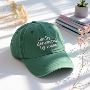 May include: A green baseball cap with the embroidered text "easily distracted by rocks" in white. The cap has a curved brim and a fabric strap closure. The hat is made of a soft, durable material.
