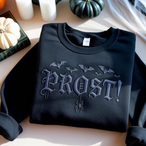 May include: Black sweatshirt with embroidered gray bats and the word "PROST!" in gothic lettering. A spider hangs below the word. The sweatshirt is a Halloween-themed apparel item.