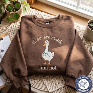 Puede incluir: Una sudadera marrón de cuello redondo con el bordado de una oca blanca que lleva un gorro de enfermera y un estetoscopio azul. El texto sobre la oca dice vitals are stable y el texto debajo dice i am not.