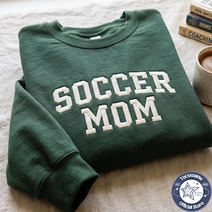 May include: A dark green crewneck sweatshirt featuring the text SOCCER MOM embroidered in large, white, block-style letters across the chest. The garment has long sleeves with ribbed cuffs and a ribbed neckline, showcasing a clean, simple design.