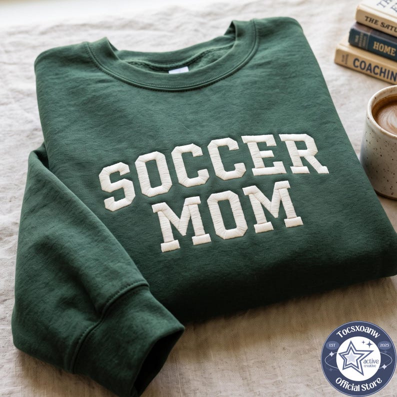 May include: A dark green crewneck sweatshirt featuring the text SOCCER MOM embroidered in large, white, block-style letters across the chest. The garment has long sleeves with ribbed cuffs and a ribbed neckline, showcasing a clean, simple design.