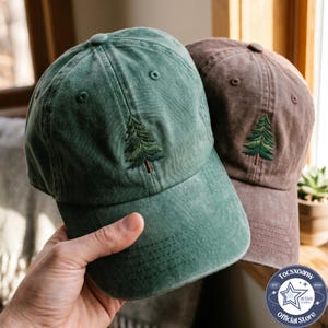 May include: A green baseball cap and a brown baseball cap, both featuring an embroidered evergreen tree design on the front panel. Each cap has a structured crown, a curved brim, and stitched eyelets for ventilation on the upper panels.