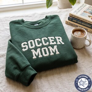 May include: A dark green crewneck sweatshirt featuring the white embroidered text SOCCER MOM in a bold block font across the chest. The garment has long sleeves, a ribbed collar, and a soft fabric texture, presented as a folded piece of apparel.