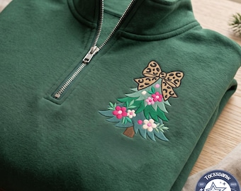 Può includere: Una felpa verde scuro con mezza zip e un ricamo raffigurante un albero di Natale stilizzato decorato con fiori rosa e bianchi, sormontato da un grande fiocco con stampa leopardata. Il colletto presenta una chiusura a zip color argento.