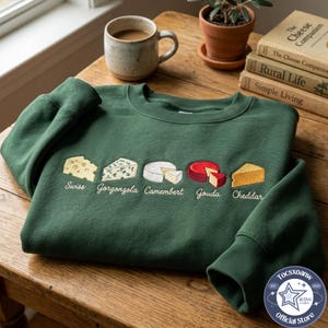 May include: A dark green crewneck sweatshirt featuring embroidered illustrations of five cheese varieties: Swiss, Gorgonzola, Camembert, Gouda, and Cheddar. Each cheese is labeled with its name in white cursive script below the respective graphic.