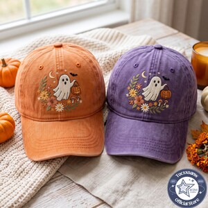 May include: Two baseball caps, one orange and one purple, each featuring an embroidered design of a white ghost holding a jack-o'-lantern, surrounded by colorful autumn flowers, mushrooms, a crescent moon, stars, and a small black bat.
