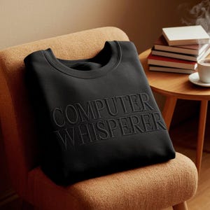 May include: A black sweatshirt with the words "COMPUTER WHISPERER" embroidered in a matching dark thread. The sweatshirt is folded and resting on a tan chair. A small wooden table with books and a cup of coffee is in the background.