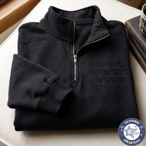 May include: A black quarter-zip sweatshirt featuring a high collar and ribbed cuffs. The chest area displays embroidered text in a dark gothic font that reads, read book, lift weights be happy. The garment has a silver-toned metal zipper pull.
