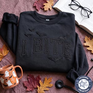 Puede incluir: Una sudadera negra de cuello redondo con el texto bordado I BITE en fuente universitaria, acentuada con murciélagos bordados. La sudadera se muestra junto a una taza con patrón de fantasmas, un libro abierto y gafas de montura negra.