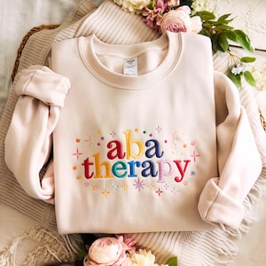 May include: A cream-coloured sweatshirt featuring the embroidered words "aba therapy" in a rainbow of colours. The design includes colourful starbursts and small dots. The sweatshirt is folded on a neutral-toned surface.