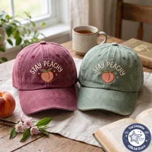 May include: Two baseball caps in maroon and sage green, each featuring an embroidered peach graphic and the text "STAY PEACHY" in white. The caps have a structured crown, a curved brim, and a button on top, showcasing a washed fabric texture.