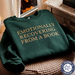 Puede incluir: Una sudadera de cuello redondo en color verde bosque oscuro con el texto bordado EMOTIONALLY RECOVERING FROM A BOOK en hilo dorado. La prenda tiene mangas largas, un cuello acanalado y un dise&ntilde;o cl&aacute;sico de su&eacute;ter con textura de tela suave.