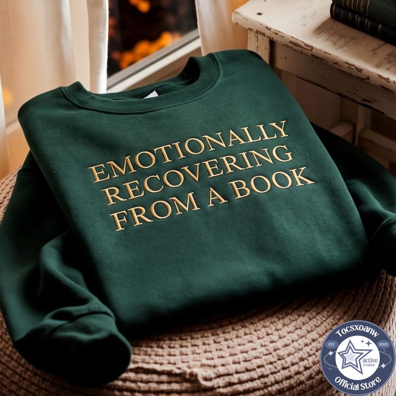 Puede incluir: Una sudadera de cuello redondo en color verde bosque oscuro con el texto bordado EMOTIONALLY RECOVERING FROM A BOOK en hilo dorado. La prenda tiene mangas largas, un cuello acanalado y un dise&ntilde;o cl&aacute;sico de su&eacute;ter con textura de tela suave.