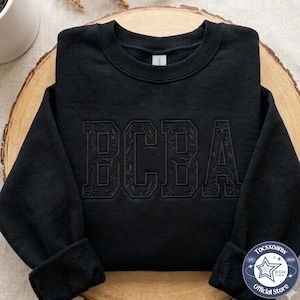 以下が含まれることがあります： 黒のクルーネックトレーナー。黒い糸でBCBAの文字が刺繍されています。文字はブロック体で、それぞれの文字の内側には複雑な花と蔓の模様がステッチで施されています。