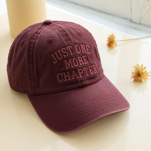 May include: A maroon baseball cap with the embroidered text "JUST ONE MORE CHAPTER" in a matching color. The cap has a curved brim and a button on top. The hat is a solid color and appears to be made of a soft fabric.