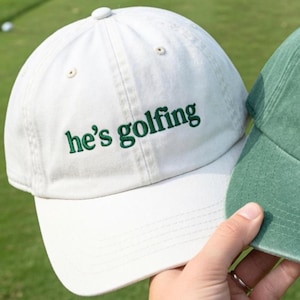 May include: A white baseball cap with the embroidered text "he's golfing" in green. The cap has a curved brim and a button on top. A second green cap is partially visible.