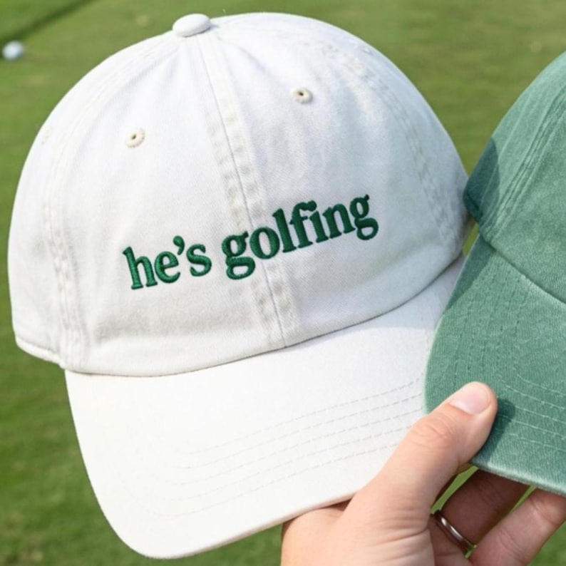 May include: A white baseball cap with the embroidered text "he's golfing" in green. The cap has a curved brim and a button on top. A second green cap is partially visible.