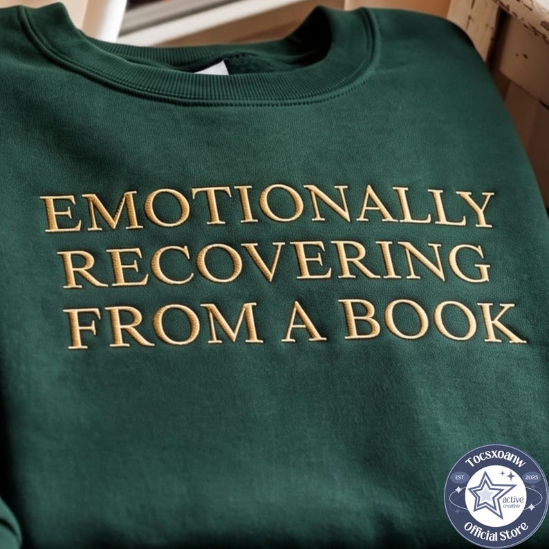 Puede incluir: Una sudadera de cuello redondo en color verde bosque oscuro con el texto bordado EMOTIONALLY RECOVERING FROM A BOOK en una fuente serif de color dorado. La prenda tiene cuello acanalado y mangas largas, con un logo circular abajo a la derecha.
