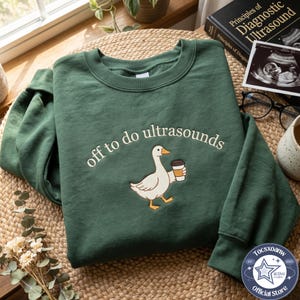May include: A dark green crewneck sweatshirt featuring an embroidered graphic of a white goose holding a coffee cup. The text "off to do ultrasounds" is arched above the goose in a cream-coloured font. The sweatshirt has long sleeves.