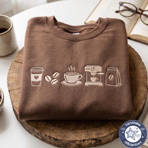 Puede incluir: Una sudadera marrón de cuello redondo con bordados de colores claros que muestran un vaso de café con un corazón, tres granos de café, una taza humeante sobre un plato, una máquina de café expreso y una bolsa de café en una línea horizontal.