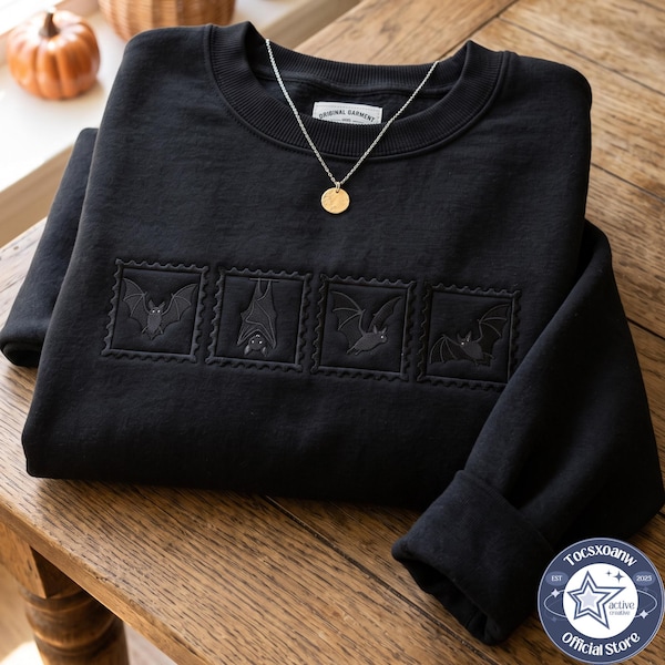 May include: A black crewneck sweatshirt featuring four embroidered square patches, each displaying a different bat illustration. The embroidery is tonal black, and the garment includes a silver-toned necklace with a round pendant resting on the collar.