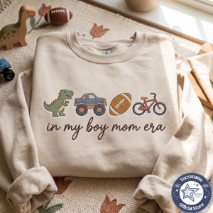 May include: A beige crewneck sweatshirt featuring a graphic print of a green dinosaur, a blue monster truck, a brown football, and a red bicycle. Below the illustrations, the text reads in my boy mom era in a dark brown cursive font.