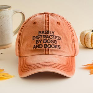 May include: A faded orange baseball cap with the embroidered text "EASILY DISTRACTED BY DOGS AND BOOKS" in black. The hat has a curved brim and a distressed look. A ceramic mug and a small pumpkin are in the background.