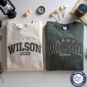 Könnte beinhalten: Zwei Rundhals-Sweatshirts, eines in Creme und eines in Olivgrün, jeweils mit dem gestickten Text CAMP WILSON EST. 2025 in einer College-Schriftart. Die Sweatshirts liegen flach und zeigen die langen Ärmel sowie die gerippten Halsausschnitte.