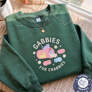 May include: A forest green crewneck sweatshirt featuring an embroidered graphic of five colorful cats and a pink pill bottle. The text reads GABBIES FOR CRABBIES in white lettering, surrounded by small white daisy flowers and four-pointed star accents.