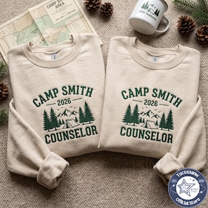 Könnte beinhalten: Zwei beige Rundhals-Sweatshirts mit dunkelgrüner Stickerei, die CAMP SMITH 2026 COUNSELOR über einer Grafik aus Tannen, Bergen und einem Zelt zeigt. Ein passender weißer Emaille-Becher mit demselben Text und Motiv ist ebenfalls zu sehen.