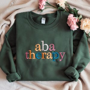 May include: A dark green sweatshirt with the embroidered text "aba therapy" in colourful letters. The letters are in orange, pink, blue, and yellow. The sweatshirt is displayed on a beige surface with pink flowers.