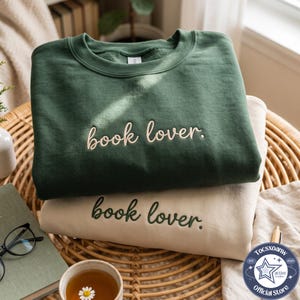 May include: A dark forest green crewneck sweatshirt with the text book lover. embroidered in cream thread, layered over a cream-coloured sweatshirt with the same text embroidered in dark green thread, both featuring a classic pullover design.